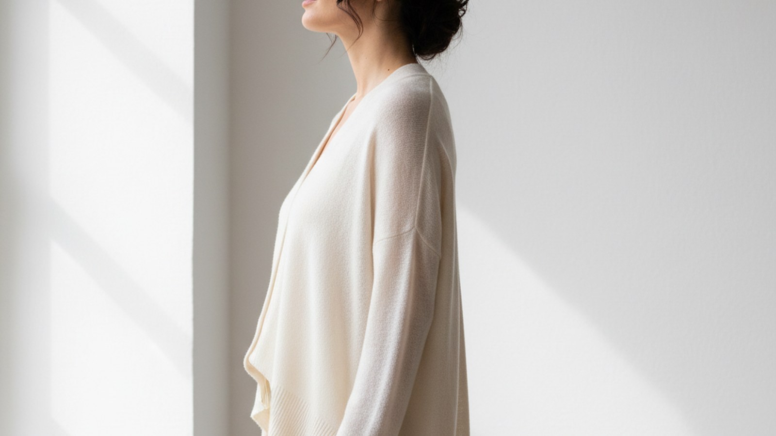 Woman in fine-gauge ivory oversized knit cardigan, dark hair loosely tucked back, standing in three-quarter profile against a