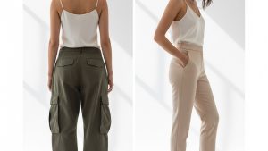 Woman in tailored trousers poses against white studio backdrop, hair loosely pulled back, face turned away from camera.