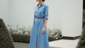 Woman in cornflower blue midi shirt dress with rolled sleeves stands in a garden under overcast daylight, face in three-quart