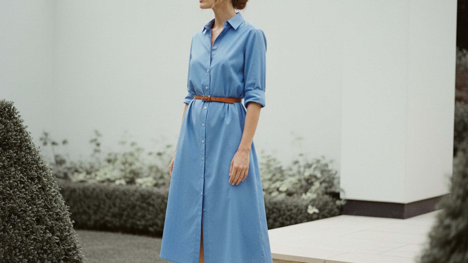 Woman in cornflower blue midi shirt dress with rolled sleeves stands in a garden under overcast daylight, face in three-quart