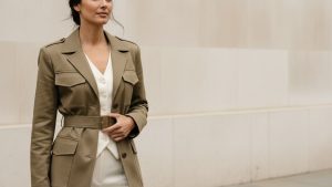 Woman in sand-khaki belted utility jacket over white silk vest and ivory wide-leg trousers, standing on a London pavement.