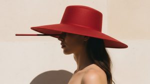 Woman in a wide-brimmed deep red hat, shot in three-quarter profile, bare shoulder visible against a pale stone wall in brigh