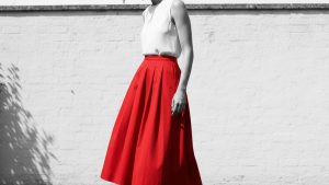 Woman in red structured midi skirt with hip pleating and dark hair, standing in a sunlit garden, weight shifted to one side.