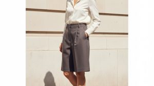 Woman mid-stride on a sunlit London pavement wearing high-waisted pleated charcoal wool-crepe shorts, one hand in pocket, chi