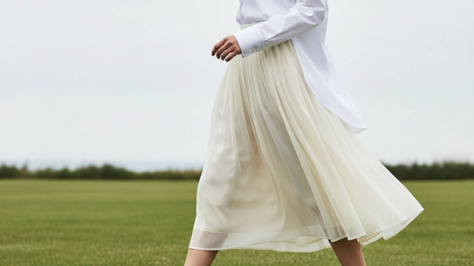 Woman mid-stride on a lawn wearing a voluminous cream organza pleated midi skirt, hem lifting slightly in the breeze.