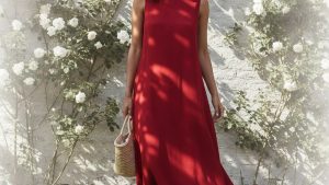 Woman with warm-toned skin and loose dark hair wearing a fluid poppy-red midi dress, standing in a sun-lit garden with white
