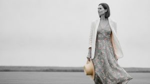 Woman in floral midi dress and open ivory linen blazer stands on a green country lawn, gazing off-camera.