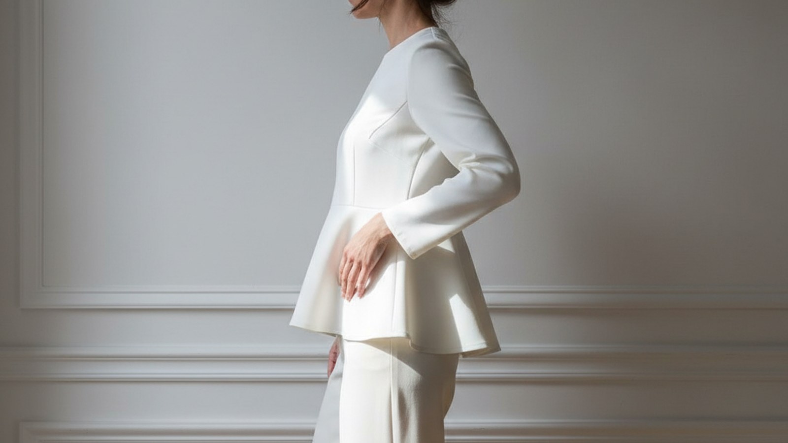 Woman in side profile wearing a chalk-white crêpe peplum top, dark hair in a low knot, one hand resting at her waist.