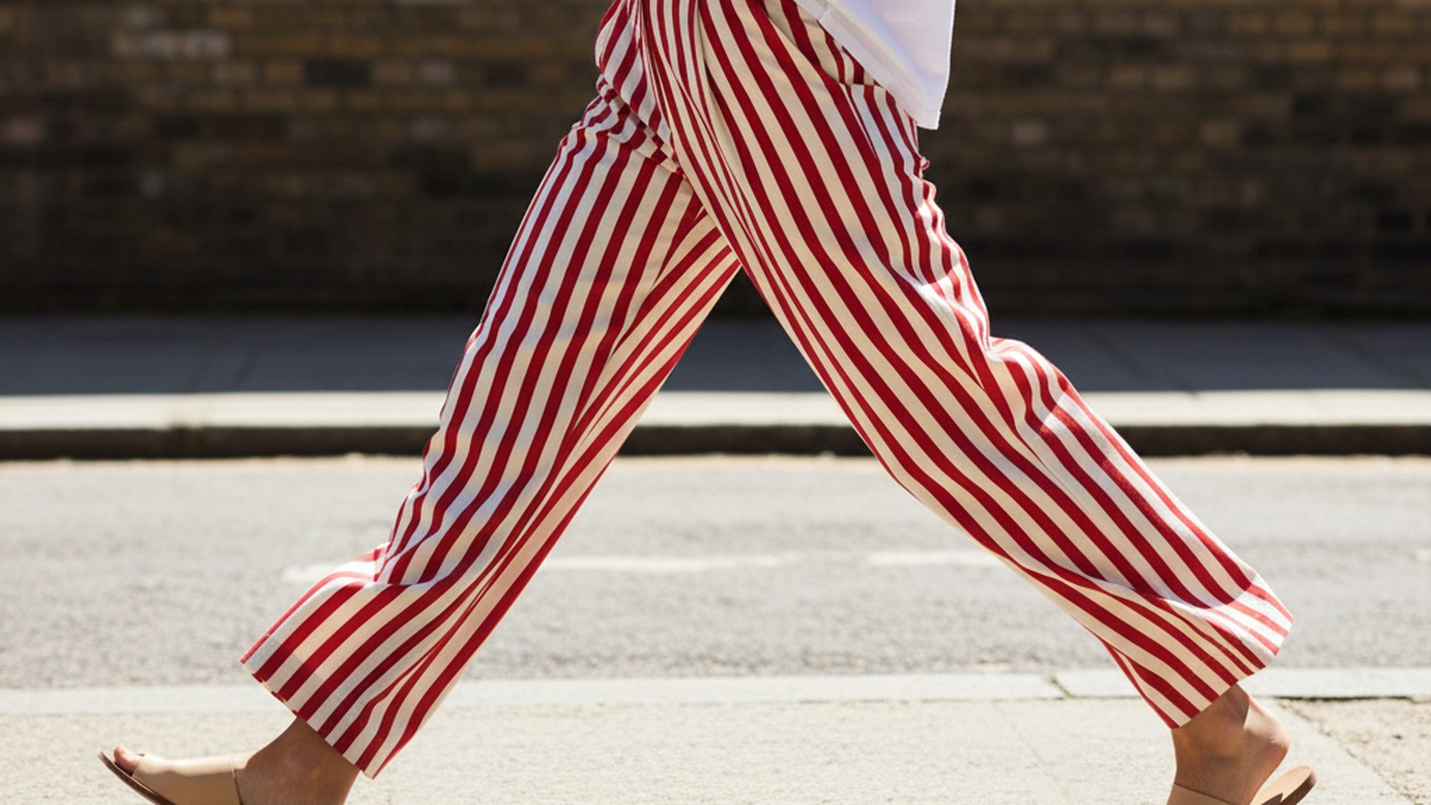 Woman mid-stride wearing wide-leg candy-striped cream and red trousers, fabric swaying in motion, on a sun-bleached pavement.