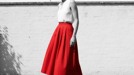 Woman in red structured midi skirt with hip pleating and dark hair, standing in a sunlit garden, weight shifted to one side.