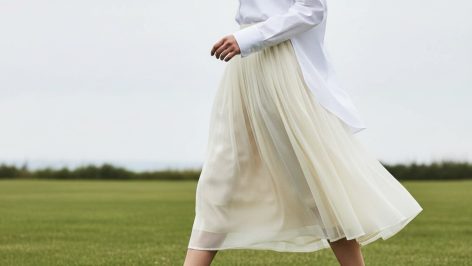 Woman mid-stride on a lawn wearing a voluminous cream organza pleated midi skirt, hem lifting slightly in the breeze.