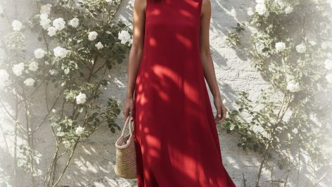 Woman with warm-toned skin and loose dark hair wearing a fluid poppy-red midi dress, standing in a sun-lit garden with white