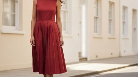 Woman in a tomato-red cotton drop-waist midi dress with pleated skirt and nude court shoes on a quiet London street.