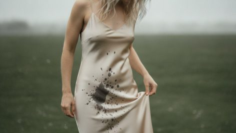 Silk vs Viscose vs Cupro: Three Home Tests That Reveal the Truth Woman lifting hem of champagne slip dress above wet grass, examining the fluid fabric with a calm downward gaze.