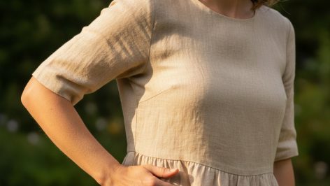 Woman smoothing a crease in an ecru linen dress with her right hand, shot from waist to shoulder in warm natural light.