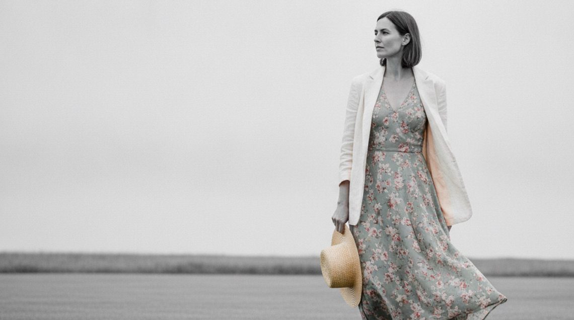 What to Wear Over a Summer Dress in the UK: The 3-Piece Capsule Woman in floral midi dress and open ivory linen blazer stands on a green country lawn, gazing off-camera.